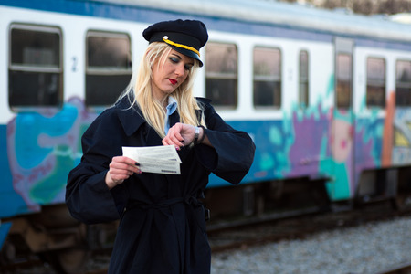 The blond hair female railroad employee or railway worker is checking time on her watch and holding two tickets, waiting for train.の写真素材