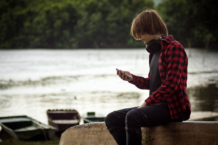 The teenage boy is using phone outdoors at waterside.の写真素材