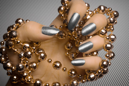 Female hand with gray nails is holding golden pearl jewelry on gray striped background, nail care and manicure concept.の写真素材