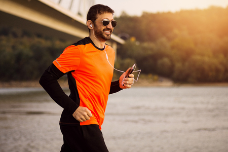 The smiling sporty man with sunglasses and earphones is jogging outdoors at the riverside.の写真素材