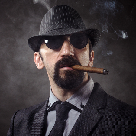 Portrait of old-fashioned gentleman in elegant suit with hat and sunglasses, smoking cigar on smoky dark gray background.の写真素材