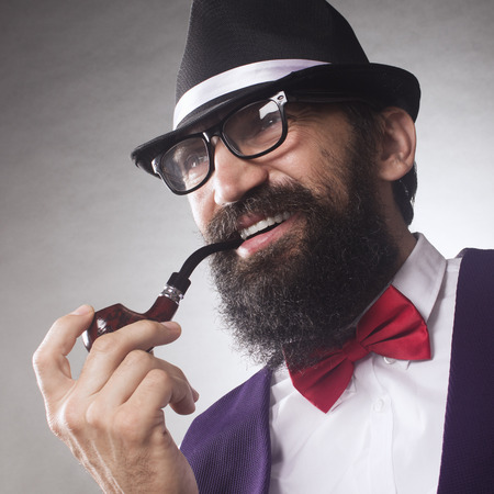 Portrait of elegant smiling bearded middle aged man with smoking pipe, eyeglasses and hat.
