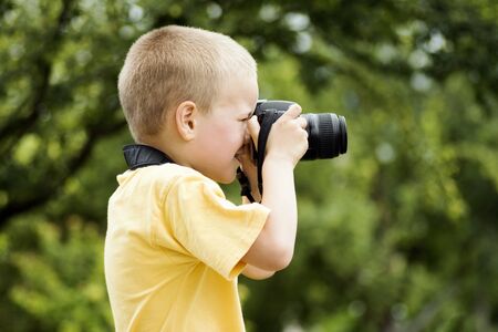 The little blond hair boy photographer is photographing outdoors on green background.の写真素材