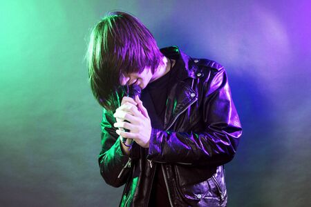 The young guy in leather jacket is singing rock music on illuminated stage on concert.