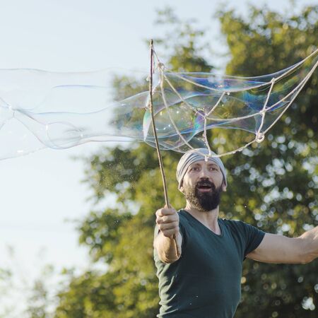 The childish playful bearded man is making giant soap bubbles outdoors.