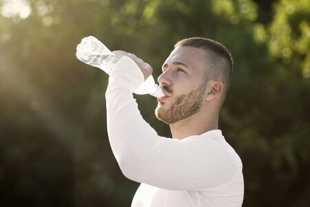 The sporty young guy is drinking water from bottle for hydrating outdoors in nature after recreation.の写真素材