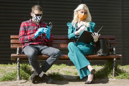 People`s life in season of coronavirus, man and woman with protective mask and gloves are sitting on bench outdoors.の写真素材