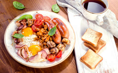 Full english breakfast with fried egg, beans, tomatoes, mushrooms, bacon, sausage, toasts and cup of...