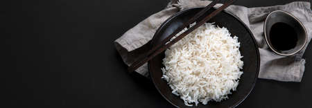 Boiled white asian-style rice in black bowl with chopsticks and soy sauce on the side. Dark background. Top view, copy space, panorama bannerの写真素材