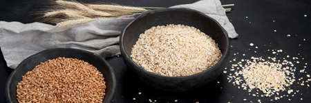 Rolled oat flakes in black bowls and golden wheat ears on dark background. Panoramaの写真素材