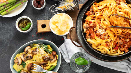 Table of italian meals on plates pasta Bolognese, ravioli, penne with chicken, sauces, parmesan and water on dark background. top view.の写真素材