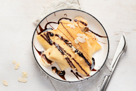 Crepe with banana, chopped almonds and chocolate sauce on white plate, table top view on a white background. Delicious french style crepes.の写真素材