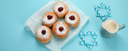 Hanukkah sweet donuts sufganiyot (traditional donuts) with fruit jelly jam and white candles on blue paper background. Jewish holiday Hanukkah concept. Panorama with copy space.の写真素材