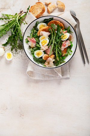 Summer fresh salad from dandelion leaves, eggs, bacon and with bread on light background. Top view, copy spaceの写真素材