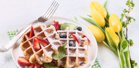 Belgian waffles sprinkled with powdered sugar with strawberries and cherries on a white background with tulips. Top view.の写真素材