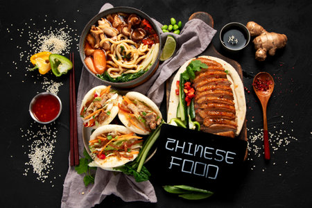 Set of Chinese dishes on dark background: sweet and sour duck, noodles soup, steamed buns. Asian style banquet or buffet, top view.の写真素材
