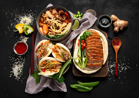 Set of Chinese dishes on dark background: sweet and sour duck, noodles soup, steamed buns. Asian style banquet or buffet, top view.の写真素材