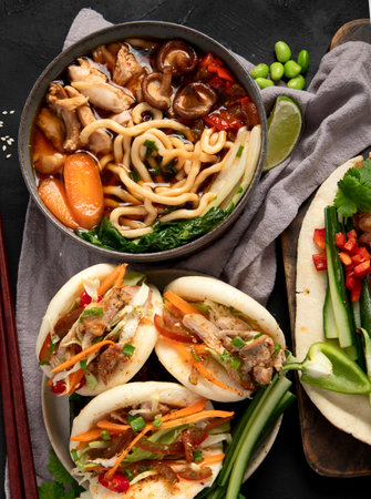 Set of Chinese dishes on dark background: sweet and sour duck, noodles soup, steamed buns. Asian style banquet or buffet, top view.の写真素材