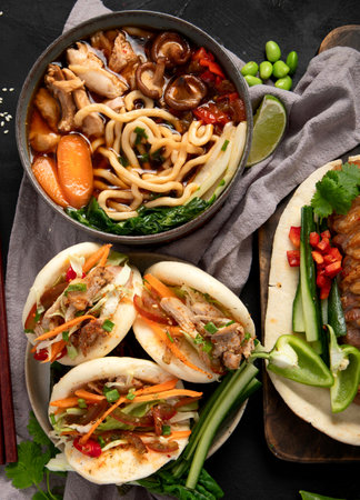 Set of Chinese dishes on dark background: sweet and sour duck, noodles soup, steamed buns. Asian style banquet or buffet, top view.の写真素材