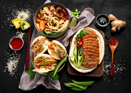 Set of Chinese dishes on dark background: sweet and sour duck, noodles soup, steamed buns. Asian style banquet or buffet, top view.の写真素材