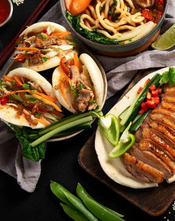 Set of Chinese dishes on dark background: sweet and sour duck, noodles soup, steamed buns. Asian...