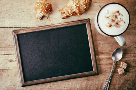 Blank blackboard with croissants, spoon and cup of coffee on wooden backgroundの写真素材