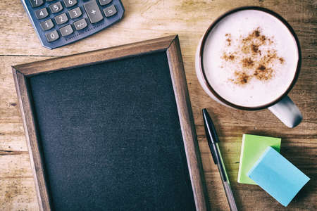 Blank blackboard with cup of coffee, pen, post-it notes and calculator on wooden backgroundの写真素材