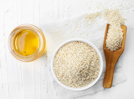 Sesame seed oil and bowl of sesame seeds on white wooden background. Top view