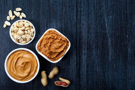 Two bowls of peanut butter and peanuts on dark wooden background from top view