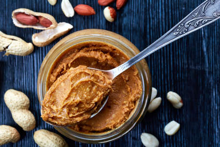 Jar and spoon of peanut butter and peanuts on dark wooden background from top viewの写真素材