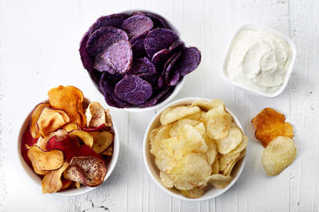 Three bowls of various vegetable chips and dip sauce on white wooden background from top view