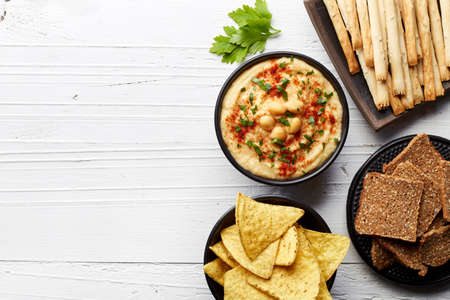 Homemade hummus and various snacks (nachos, toasts and bread sticks) on white wooden background from...