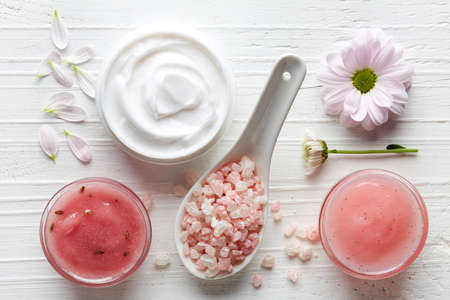 Cosmetic cream container, body scrub, bath salt and pink flowers on white wooden background from top viewの写真素材
