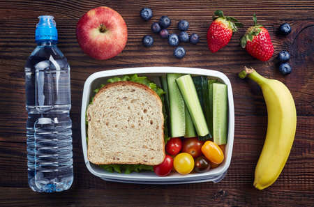 Healthy lunch box with sandwich and fresh vegetables, bottle of water and fruits on wooden...