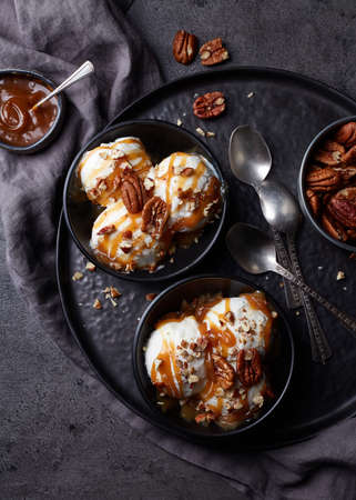 Two black bowls of vanilla ice cream balls with caramel sauce and pecan nuts on dark marble background. Top viewの写真素材