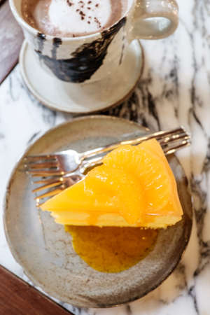 Orange cake placed on deep blue colour clay plate with small silver colour fork near a clay mug filled with hot coffee, both of them placed on marble top tableの写真素材