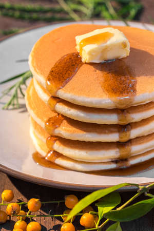 Stack of delicious pancake topped with butter and maple syrup placed on round plate with rosemary on wood table decorated with berrys and leafsの写真素材