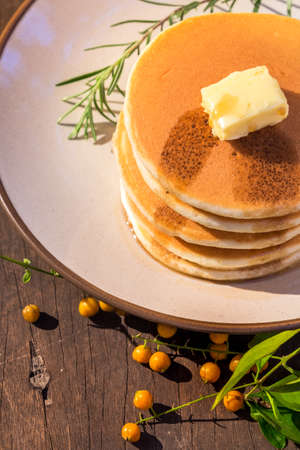 Stack of delicious pancake topped with butter and maple syrup placed on round plate with rosemary on wood table decorated with berries and leafsの写真素材