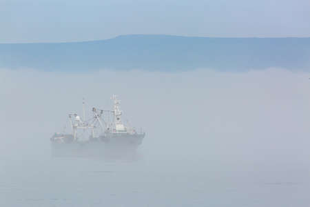 Fishing boat in thick fog on the background of mountainsの写真素材