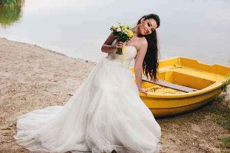 Happy bride holding a bouquet and sitting on the yellow boat on the river bank.の写真素材