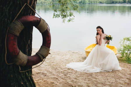 Lifebuoy on the tree and smiling bride sitting on the yellow boat on the background.の写真素材
