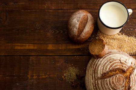 Fresh brown crispy bread, slice of bread, tasty milk in the white cup and wheat grain on the wooden tableの写真素材