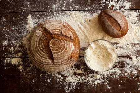 Two loaves of nice tasty brown bread and bowl of flour are on the old wooden table sprinkled with flour. Top viewの写真素材