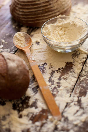 Two loaves of just baked tasty bread, bowl of flour and wooden spoon are on the brown table...
