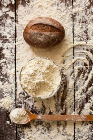 Two loaves of just baked tasty bread, bowl of flour and wooden spoon are on the brown table sprinkled with wheat flourの写真素材