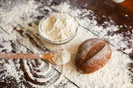 Two loaves of just baked tasty bread, bowl of flour and wooden spoon are on the brown table sprinkled with wheat flourの写真素材