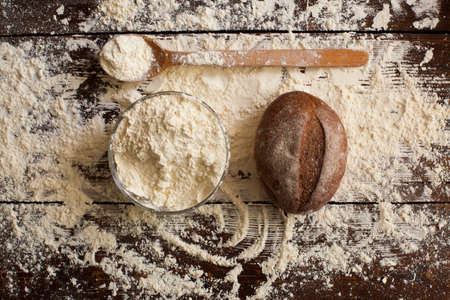 Two loaves of just baked tasty bread, bowl of flour and wooden spoon are on the brown table sprinkled with wheat flourの写真素材