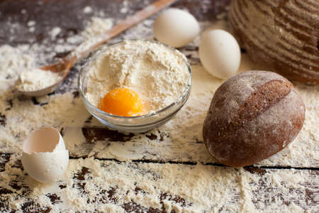 Top view of bowl of wheat flour with raw egg and wooden spoon full of flour, two loaves of nice...