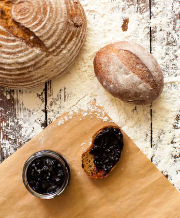 Two loaves of fresh crispy bread are on the wooden table sprinkled with flour and slice of bread with delicious berry jam from glass jar are on the cutting board.の写真素材