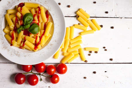 Hot delicious pasta dish with sauce on white plate prepared for dinner and raw macaroni with red cherry tomatoes on the white wooden tableの写真素材